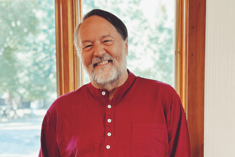 Reese Jones in a relaxed pose wearing a red shirt, standing by a bright window with soft natural lighting and outdoor scenery.