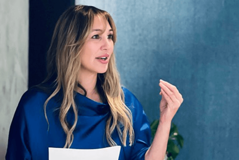 Nicole Daedone in a blue blouse giving a presentation, holding papers, focused on empowering women’s sexuality and personal growth.